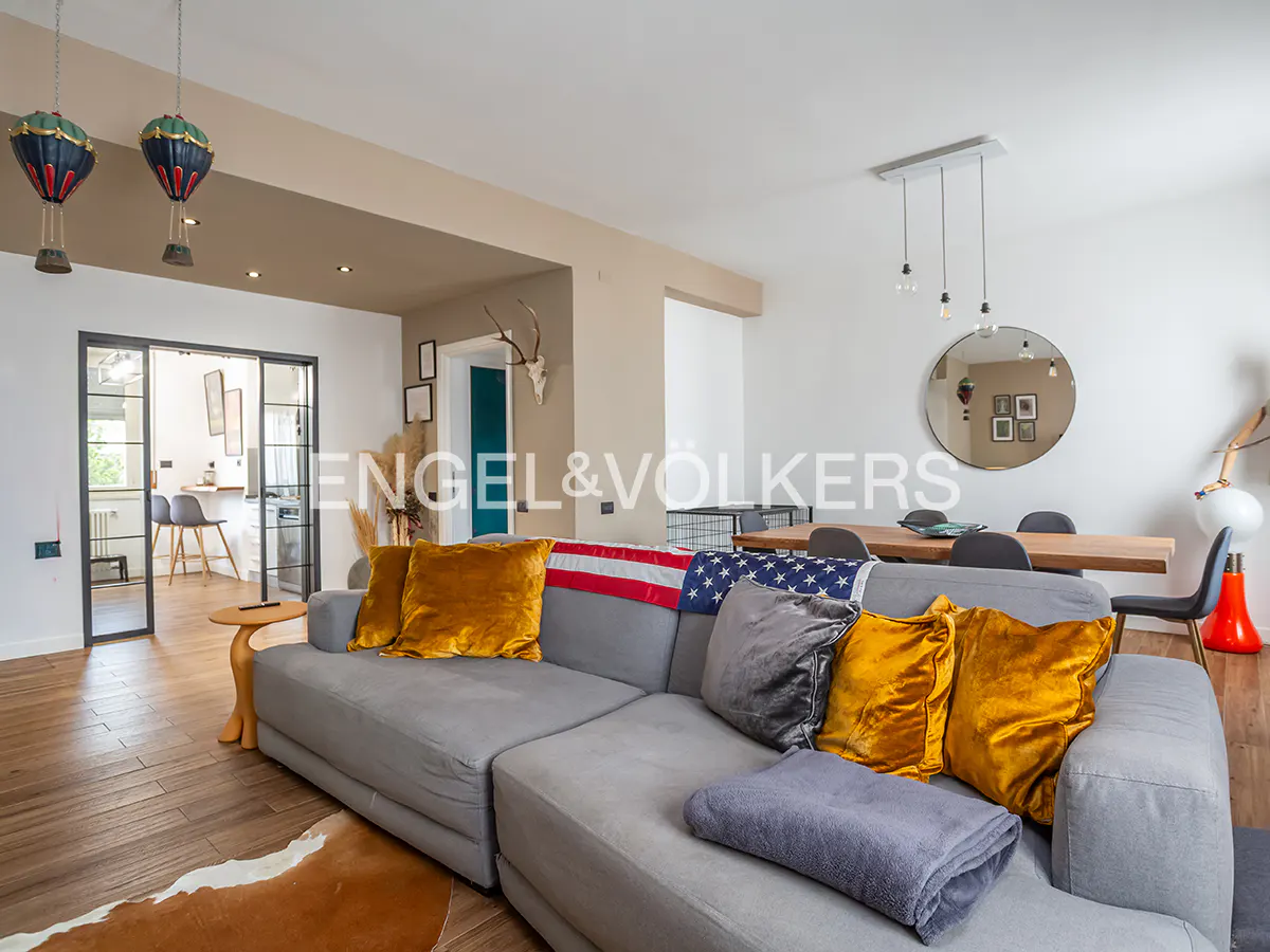 Open-concept living space with a gray sectional sofa, gold pillows, and an American flag. Dining table and kitchen visible in the background.