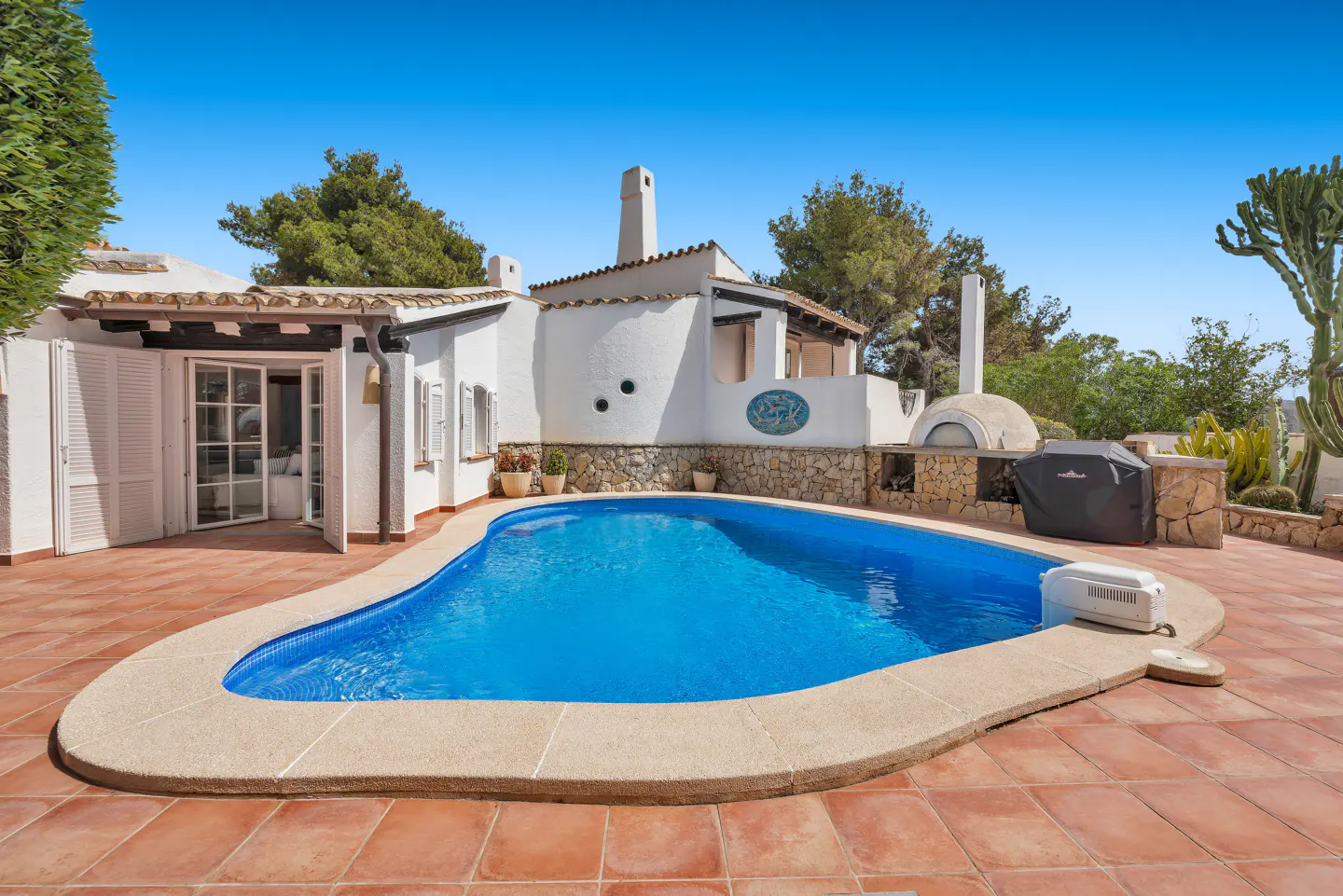 Outdoor pool with blue water, surrounded by a tan stone patio and a white house with a pizza oven.