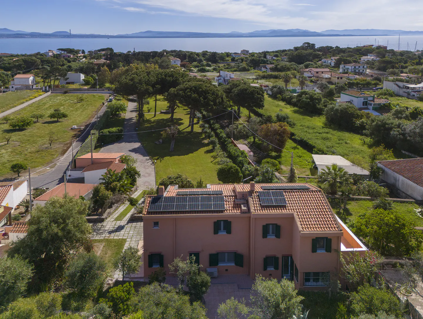 Aerial view of a peach-colored two-story house with a red tile roof and solar panels, surrounded by green trees and grass. Ocean in the background.