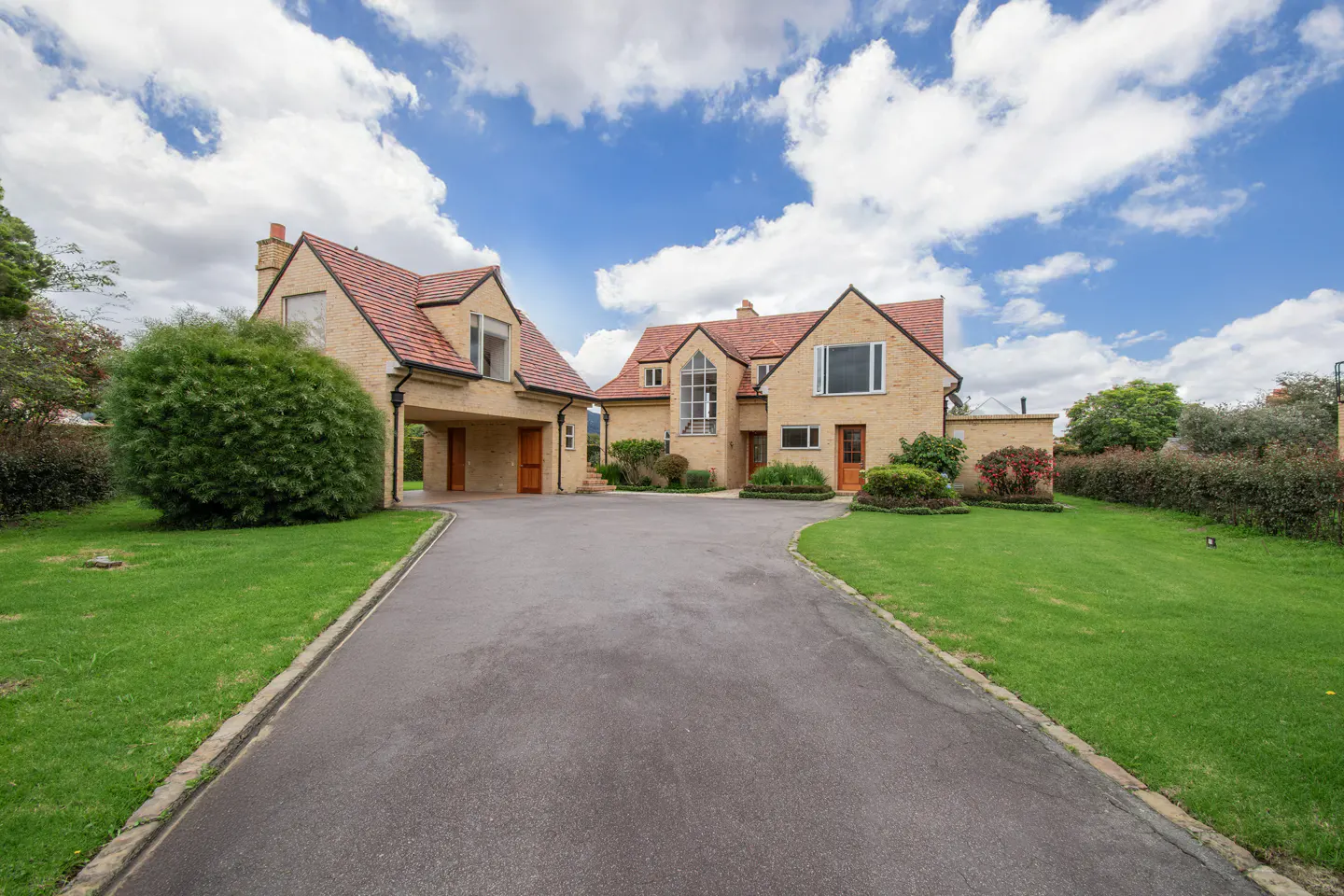 Beige brick house with a red tile roof, green lawn, and a gray driveway under a blue sky with white clouds.