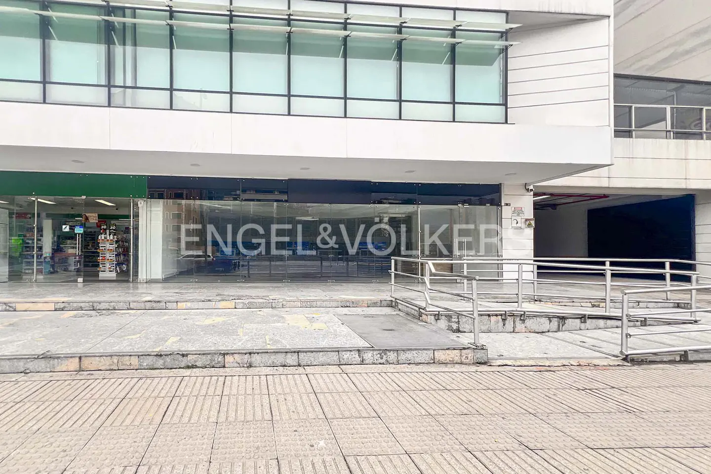 Exterior of a white building with "Engel & Völkers" on the glass storefront. A ramp leads to the entrance. A store is on the left.