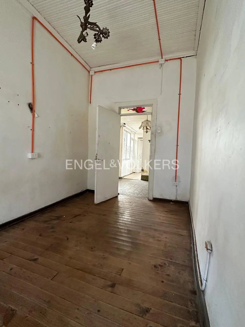 Empty room with wooden floors, white walls, exposed orange electrical conduit, and a doorway leading to another room.