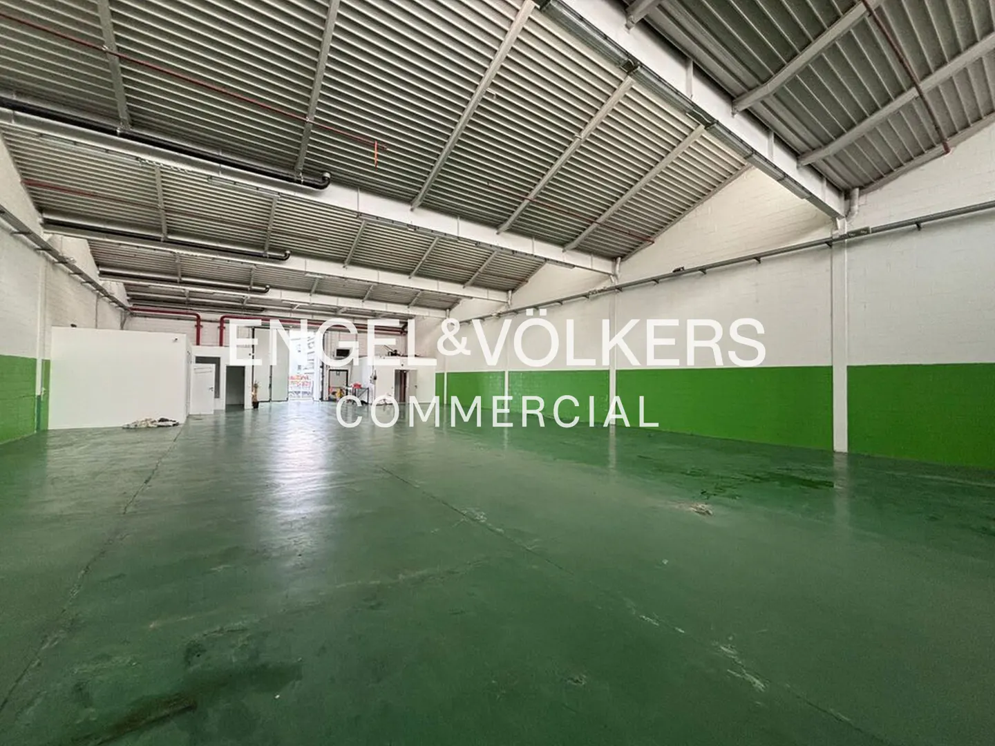 A large, empty warehouse with a green floor and white walls. The ceiling is high and metal. "Engel & Völkers Commercial" is superimposed on the image.