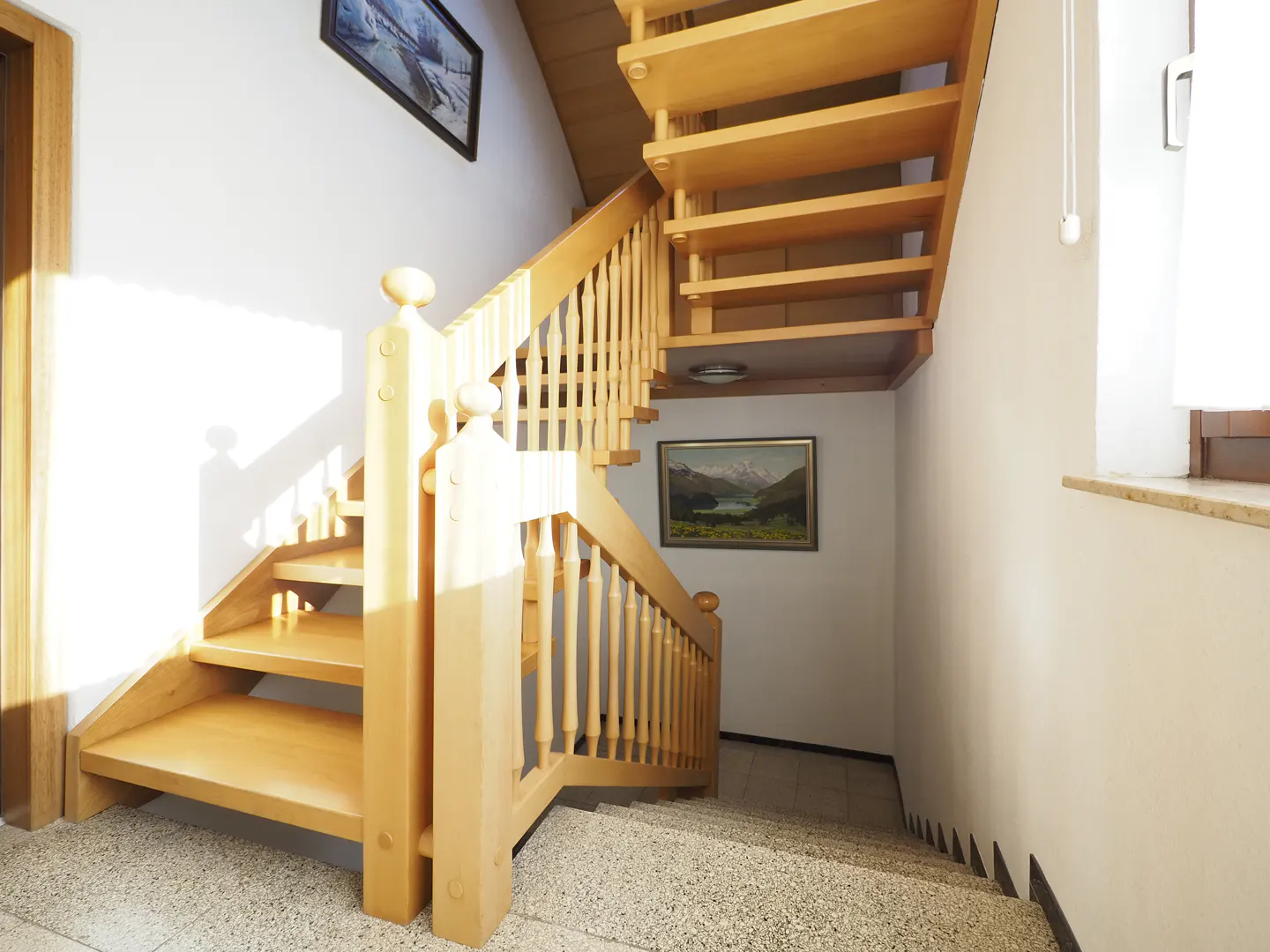 Wooden staircase with railing in a white-walled home. Artwork hangs on the walls. Natural light from a window.