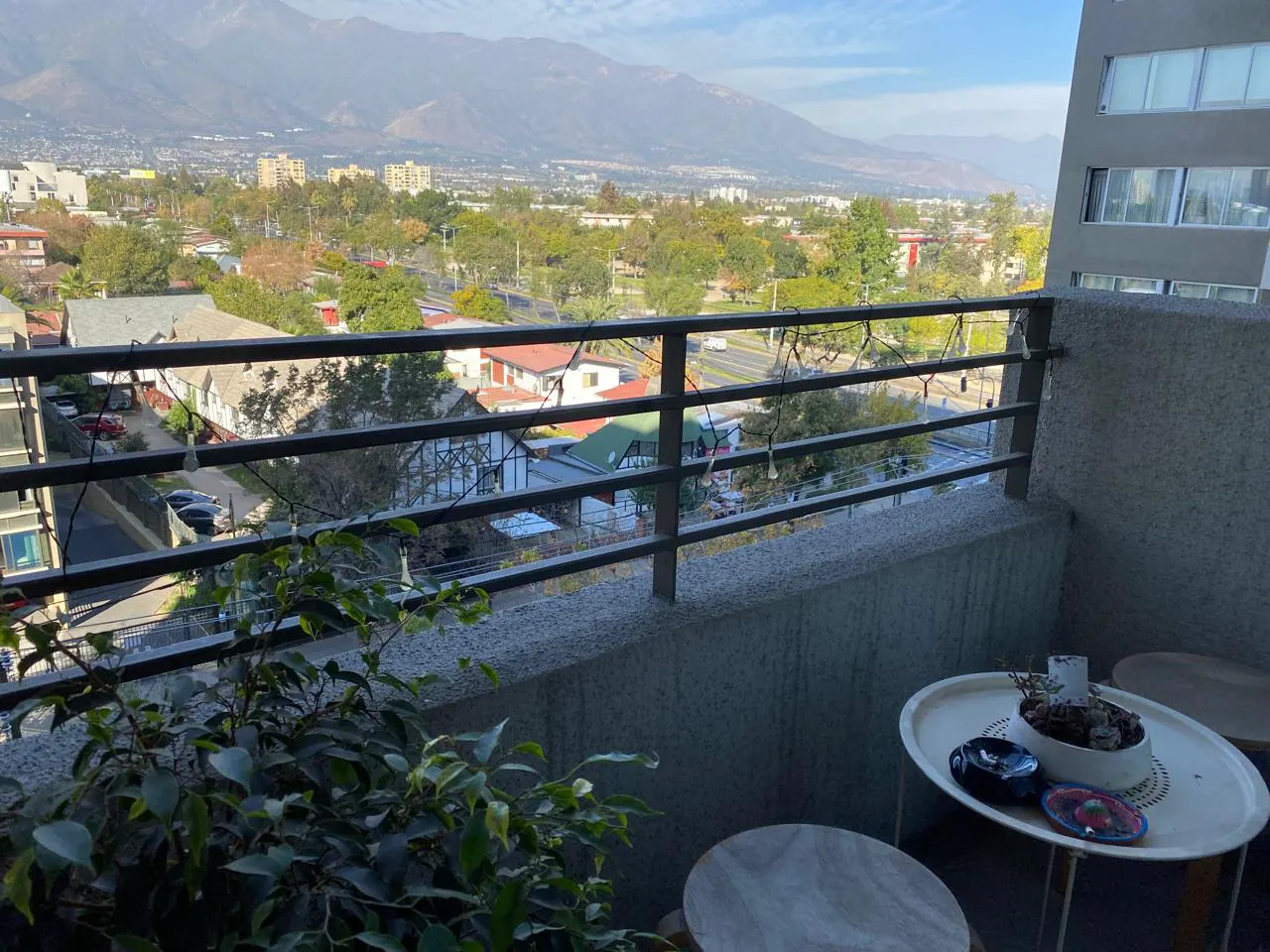 Balcony view with metal railing, plants, and small tables. Cityscape with mountains in the background.