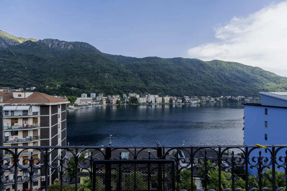 View from a balcony overlooking a calm lake, surrounded by lush green mountains and a town with buildings under a blue sky.