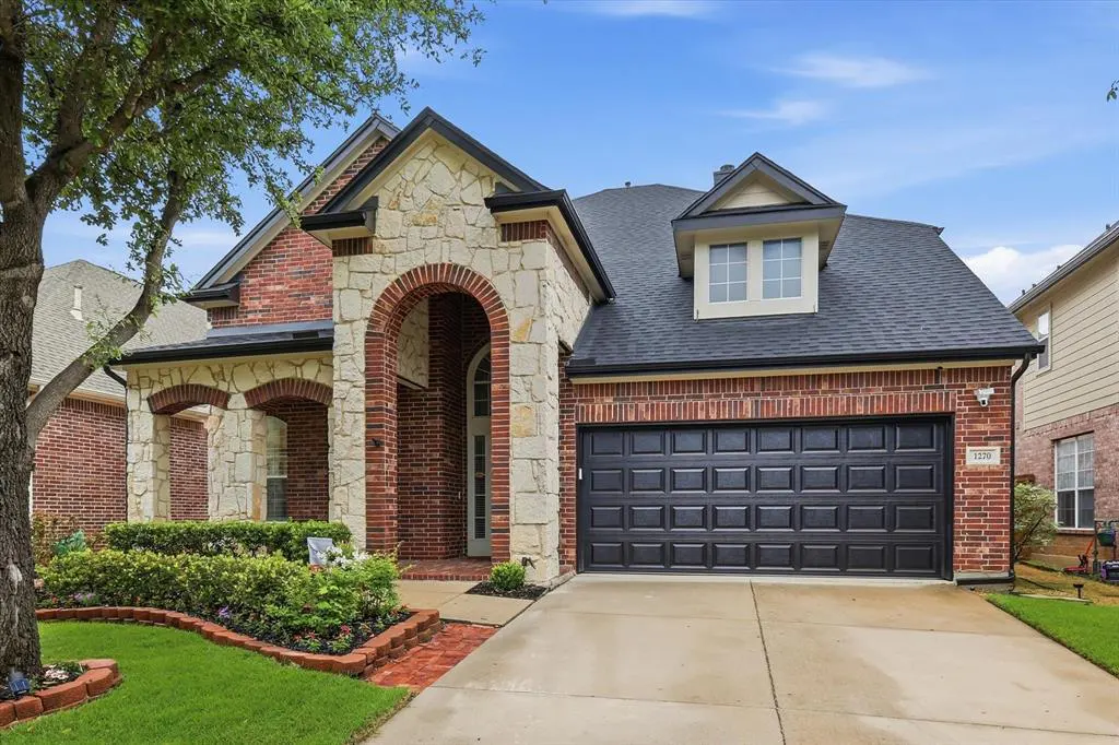 Two-story brick house with stone accents, a dark gray roof, and a two-car garage. Green lawn and landscaping in front.