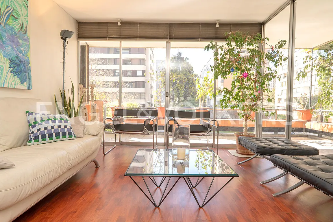 Bright living room with a white sofa, glass table, and black chairs. Large windows overlook a balcony with plants and city views. Hardwood floors.