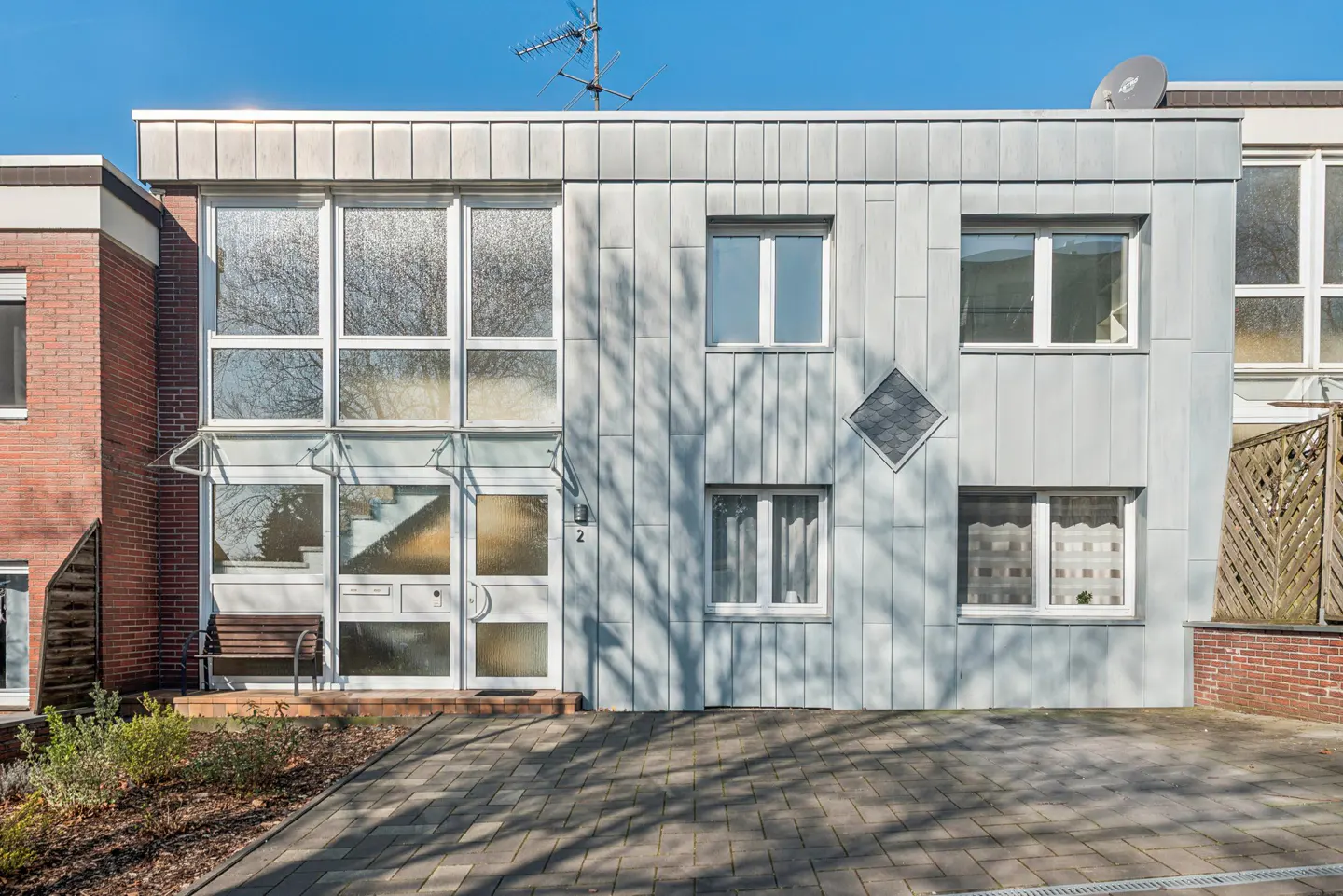Modern two-story townhouse with gray metal siding, large windows, and a brick facade on the left. A paved walkway leads to the front door.