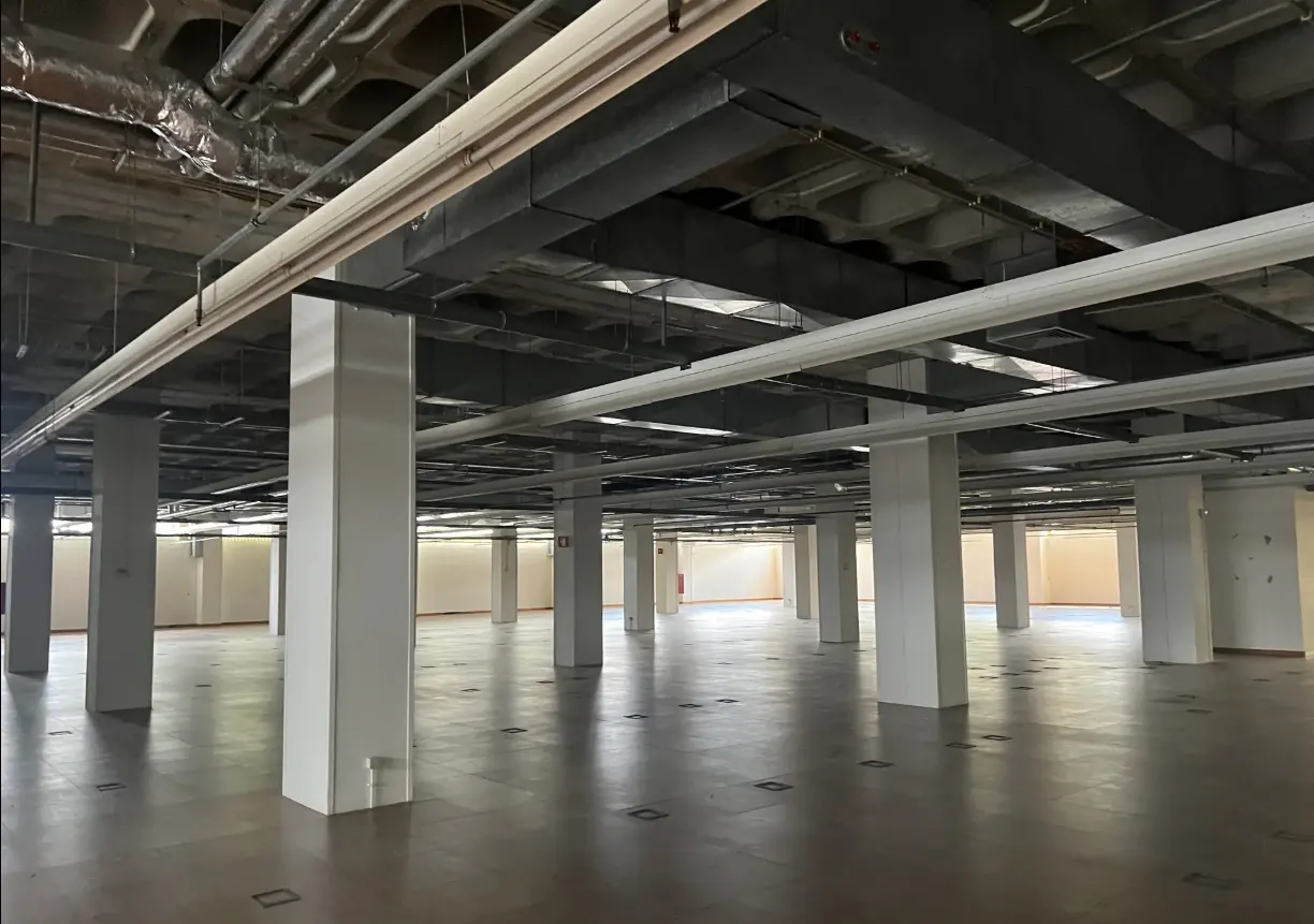 Large empty commercial space with white support columns and exposed ceiling pipes and ductwork.