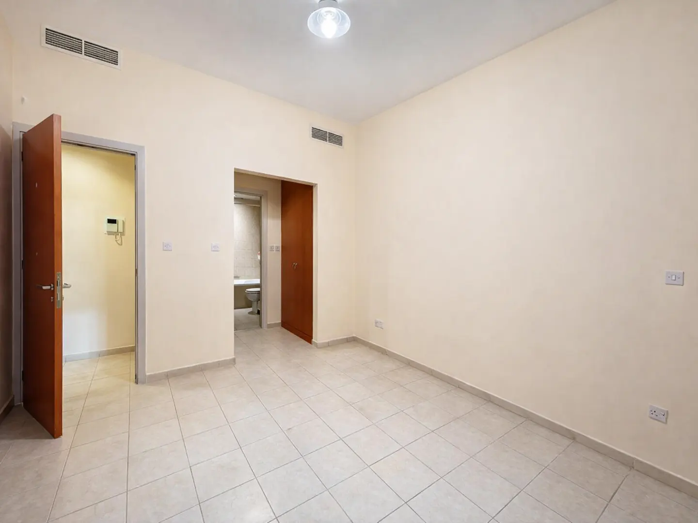 Empty room with cream walls, light tiled floors, a ceiling light, and two doorways leading to a bathroom and hallway.