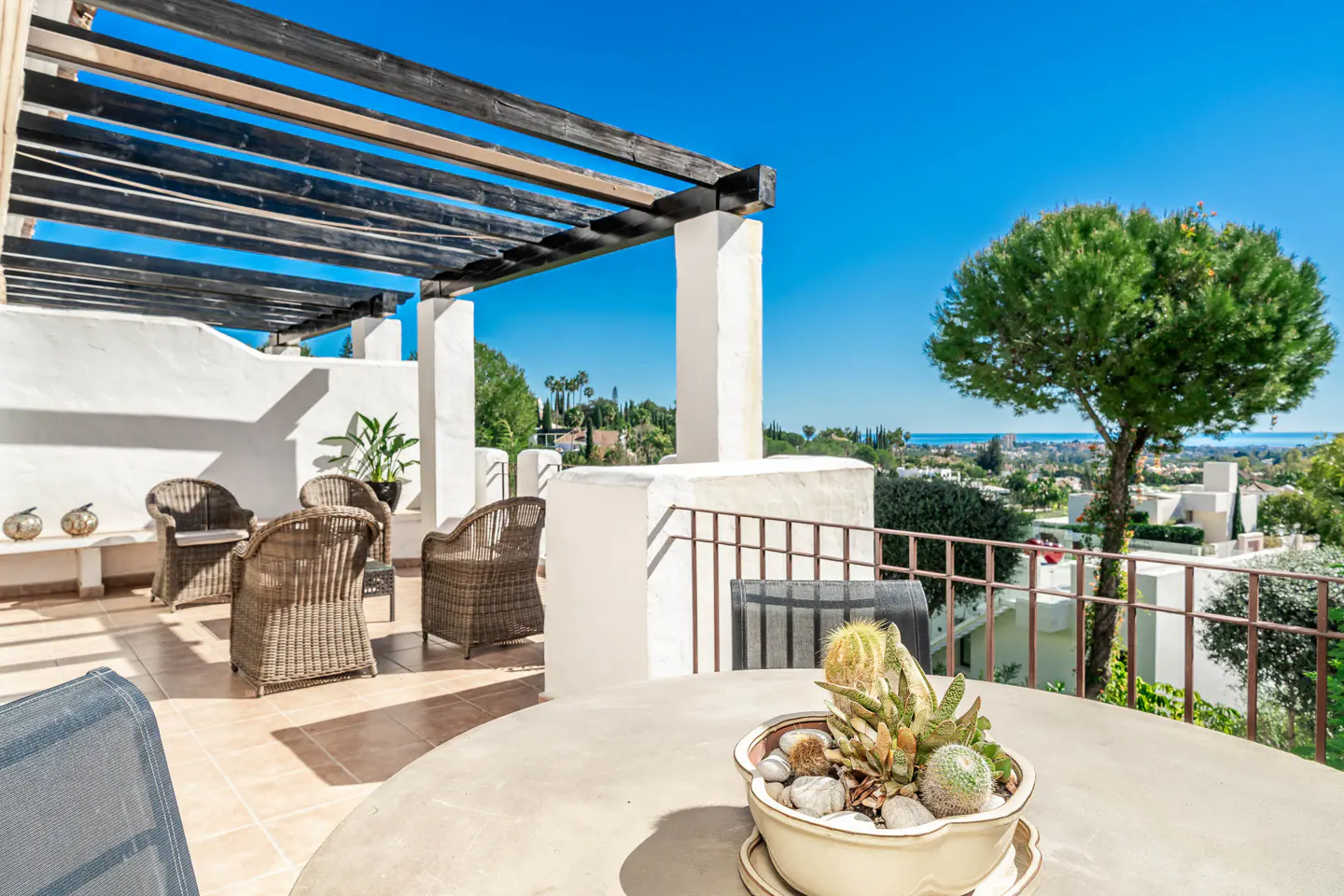 Outdoor patio with wicker chairs, a round table with a cactus planter, and a pergola, overlooking a scenic view of trees and the ocean.