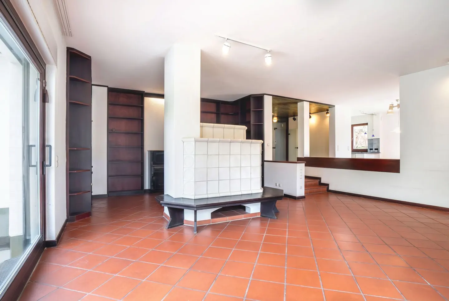 An open-concept living room with terracotta tile floors, dark wood bookshelves, and a white tiled fireplace with a bench.
