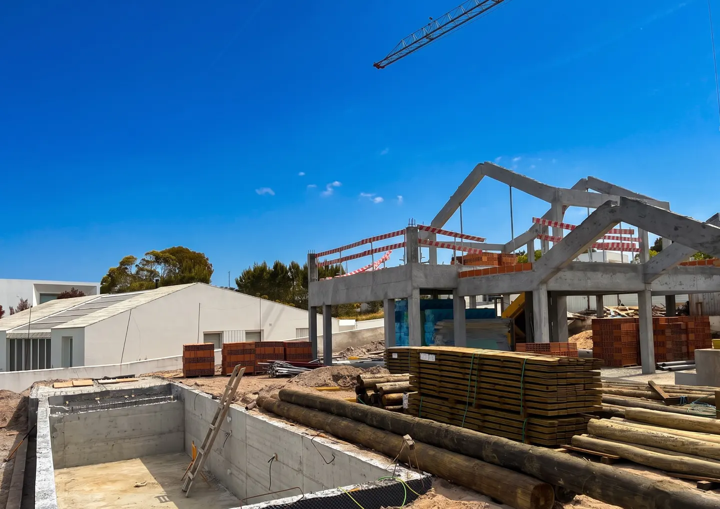 Construction site with a concrete pool, lumber, and a building frame under a blue sky. A crane is visible in the background.