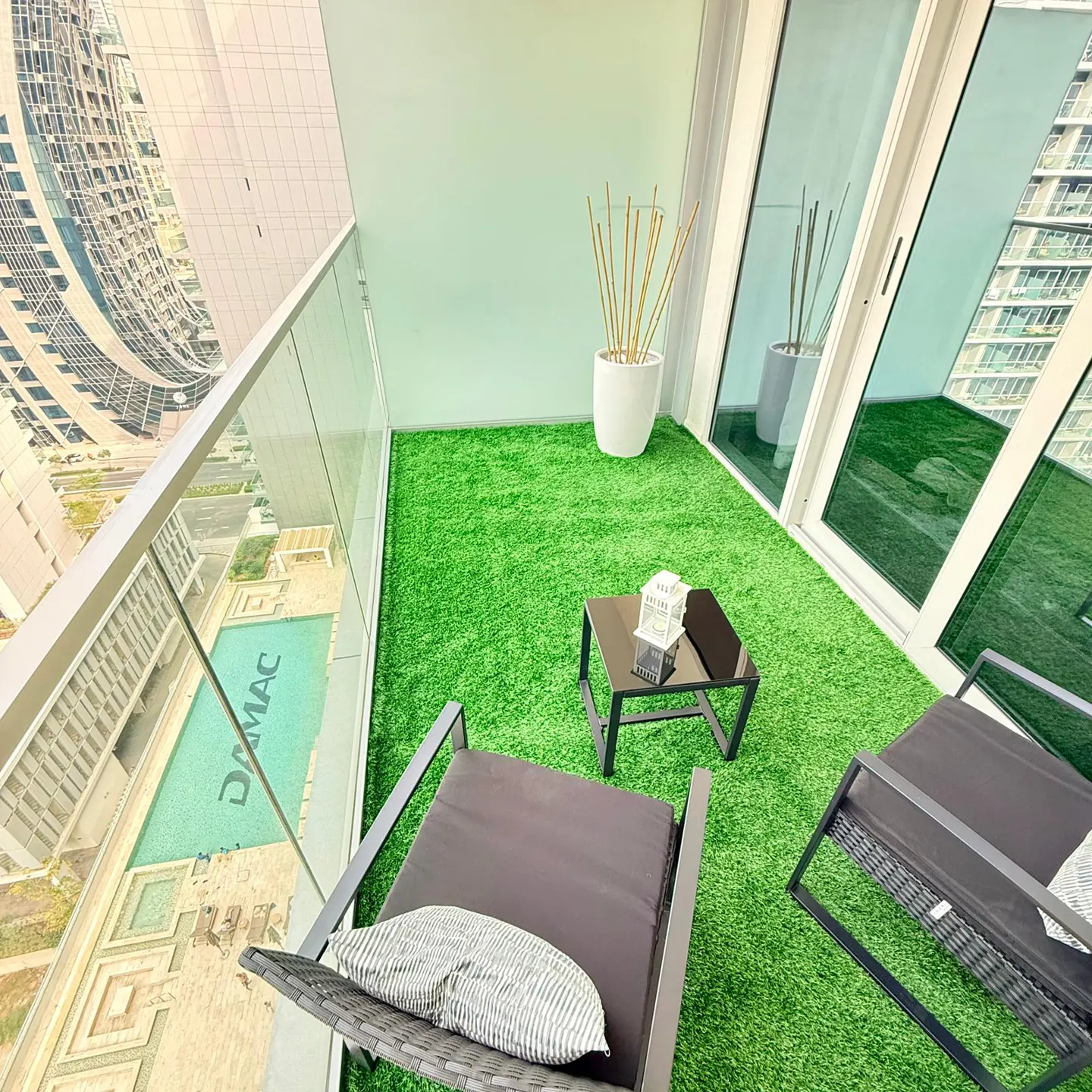 Balcony with green turf, chairs, and a small table. A white planter with bamboo is near sliding glass doors. A pool is visible below.