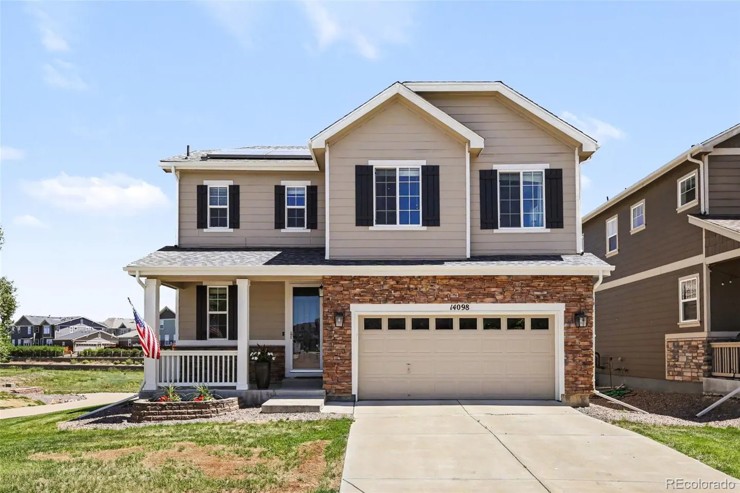 Two-story suburban home with tan siding, stone accents, black shutters, a two-car garage, and an American flag on the porch.