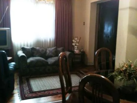 A living room with a patterned sofa, a rug, and a dining table with wooden chairs. A window with sheer curtains is visible.