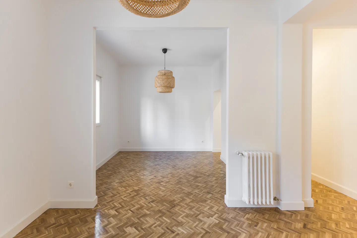 Bright, empty room with white walls, herringbone wood floors, and woven light fixtures. A white radiator is visible.