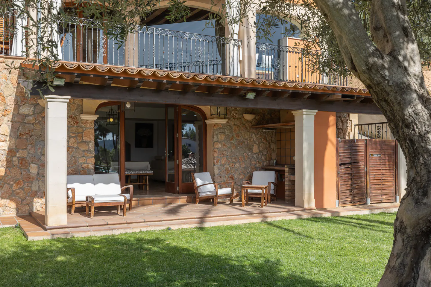 Outdoor patio with stone walls, wooden furniture, and green lawn. Balcony above with metal railing. Tree on right side.