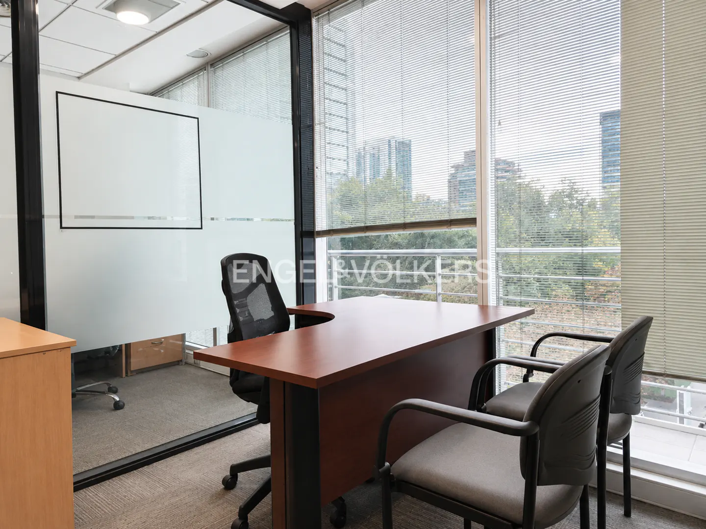 Office interior with a brown desk, two gray chairs, and a black mesh office chair. Large windows with blinds overlook a cityscape.