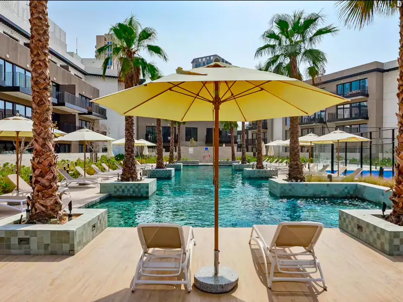 A resort pool with lounge chairs, yellow umbrellas, and palm trees. The pool is surrounded by modern buildings.