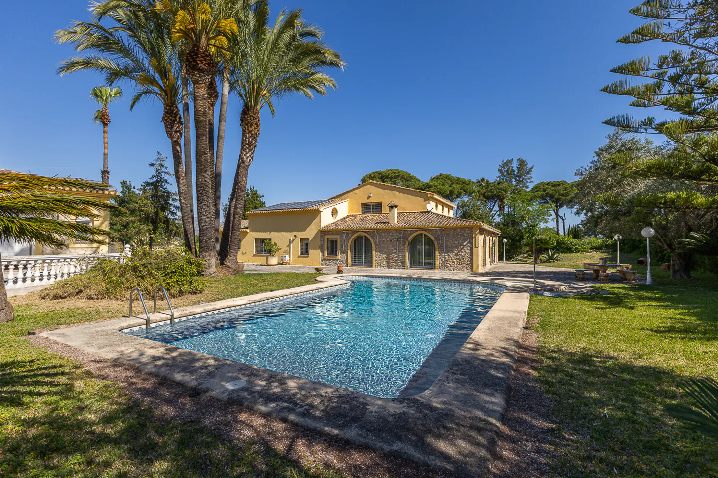 A yellow house with a stone facade overlooks a blue swimming pool surrounded by green grass and palm trees.