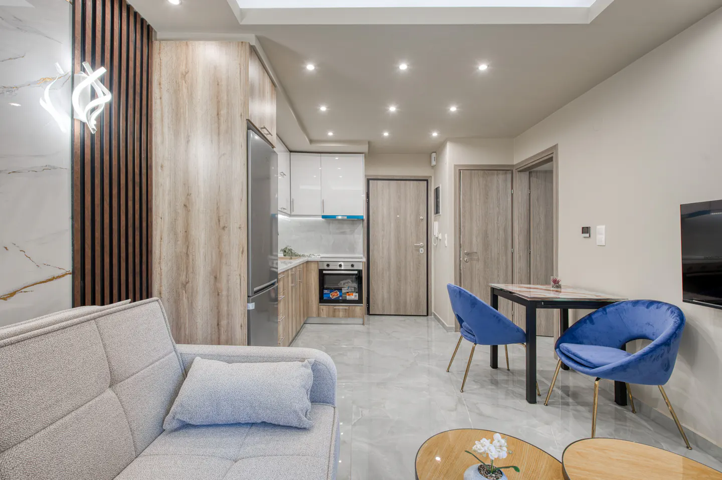 Bright, modern apartment interior with a gray sofa, blue chairs, and a compact kitchen with wood-grain cabinets.
