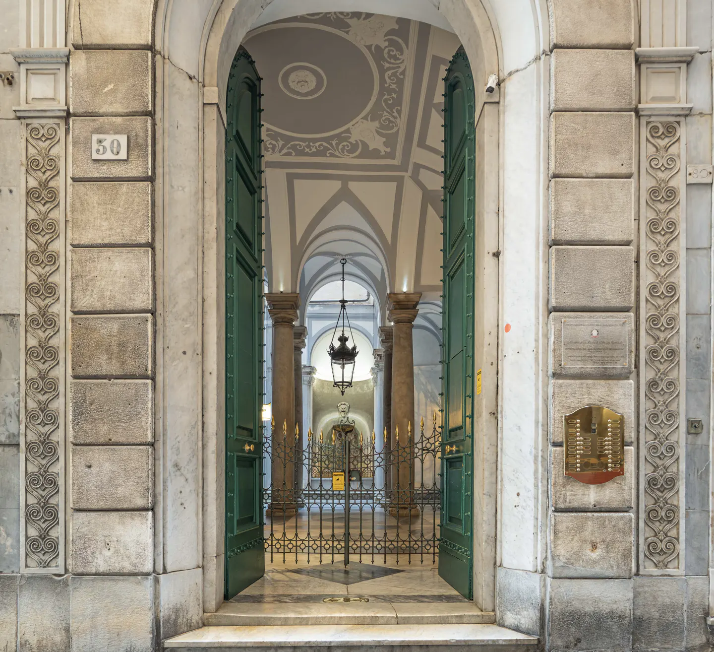 Open green doors reveal a grand hallway with columns and arches. A black lantern hangs from the ceiling. Stone walls frame the entrance.