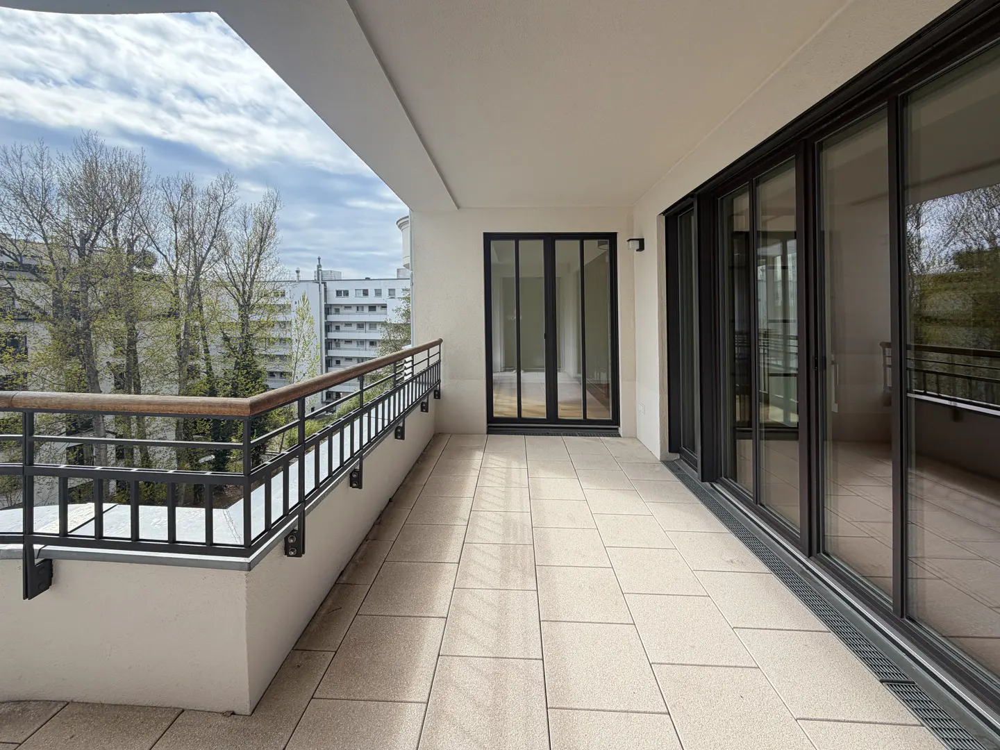 A bright, empty balcony with black metal railings and sliding glass doors. Trees and buildings are visible in the background.
