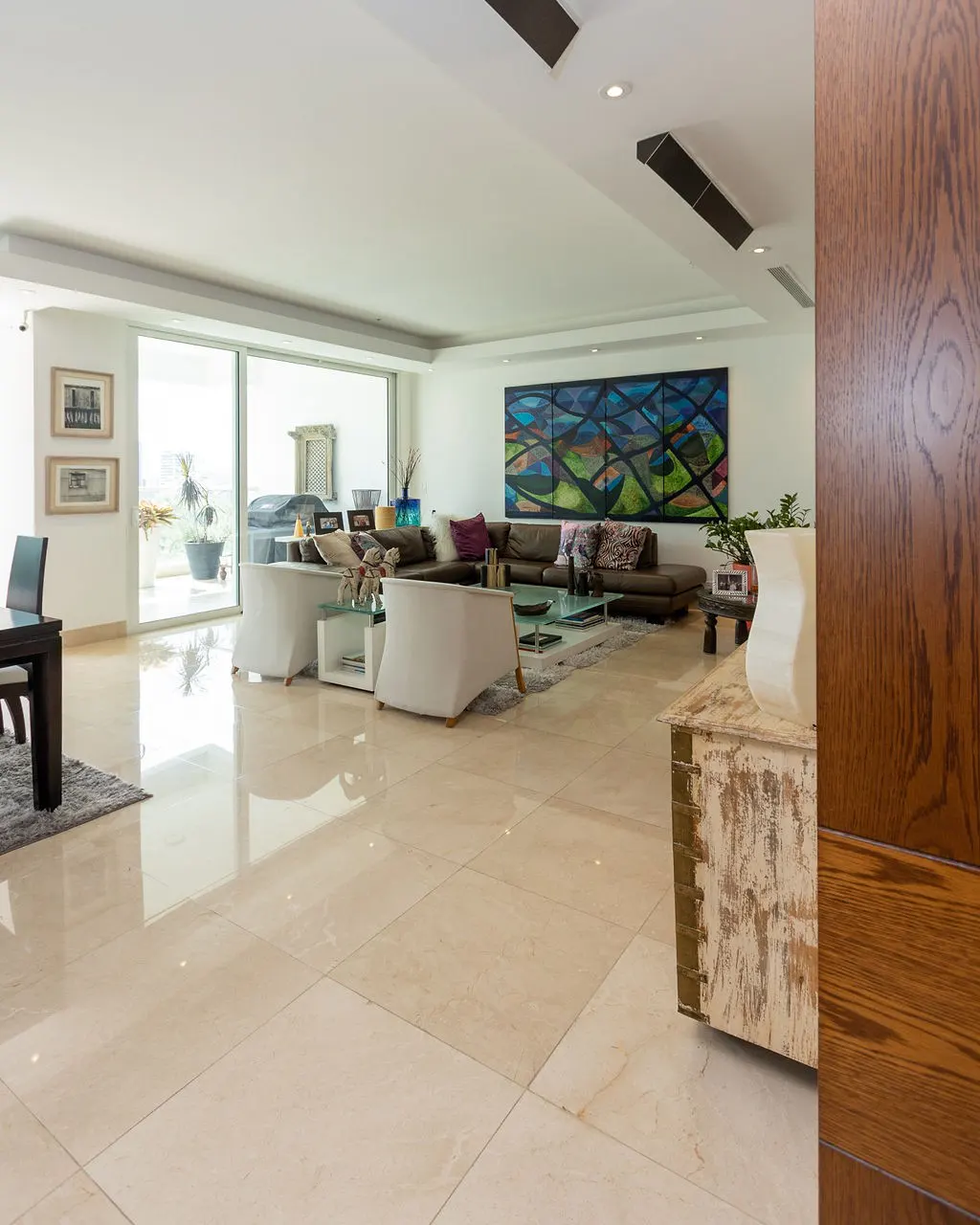 Bright, open living room with beige tile floors, white walls, and a large abstract painting. Brown sectional sofa and white armchairs.