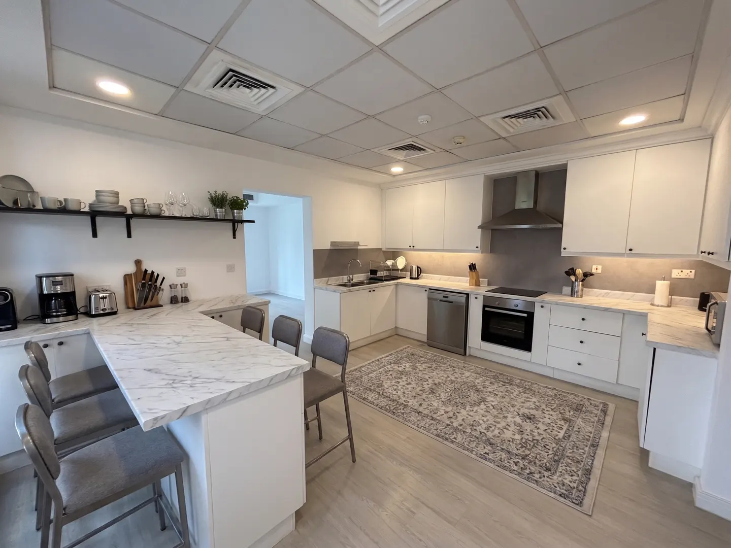 A bright, modern kitchen with white cabinets, marble countertops, and gray chairs at the breakfast bar. A patterned rug lies on the light wood floor.