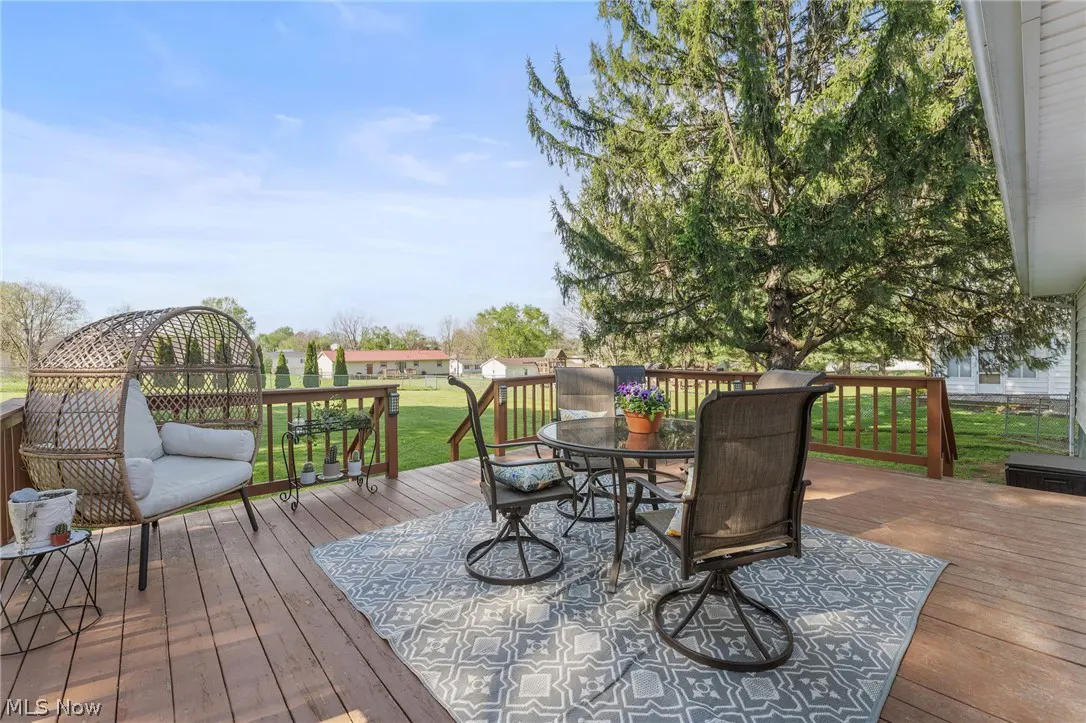 A wooden deck with a wicker chair, table, and chairs on a gray patterned rug. A large tree is in the background.