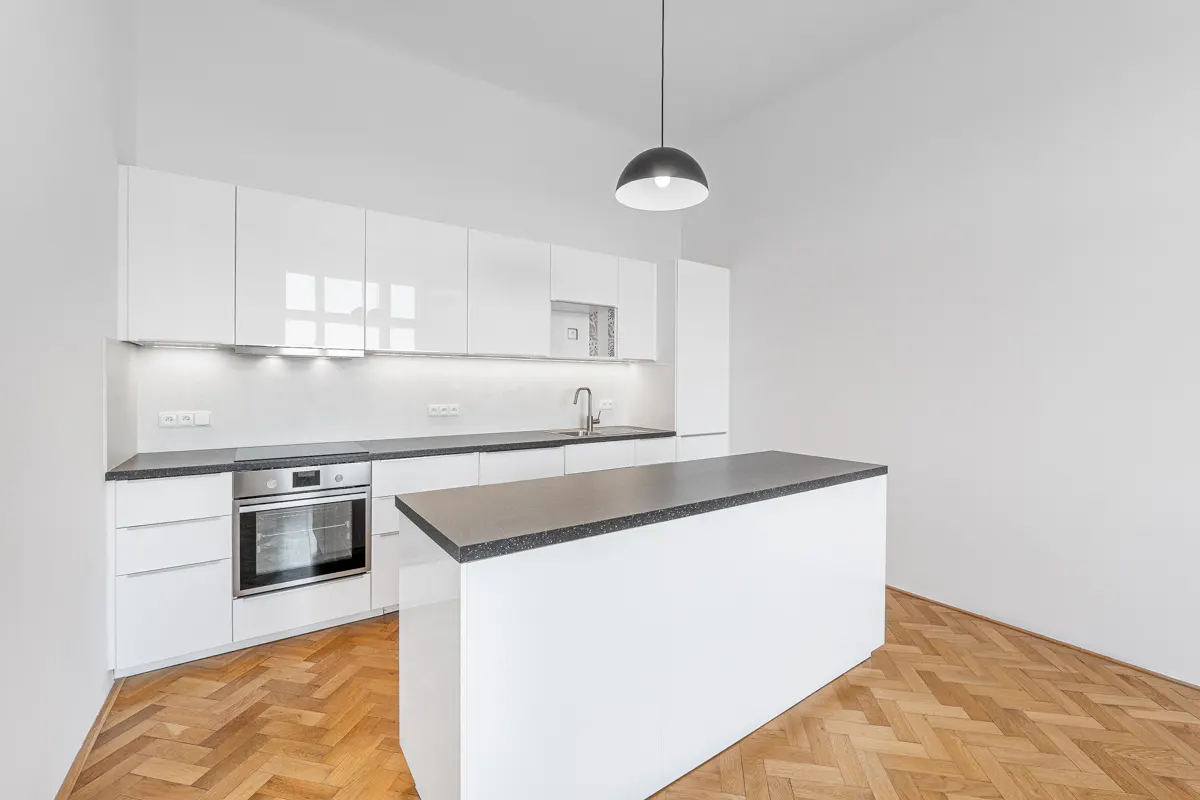 Bright kitchen with white cabinets, dark countertops, and a central island. A black pendant light hangs above the island. Herringbone wood floors add warmth.