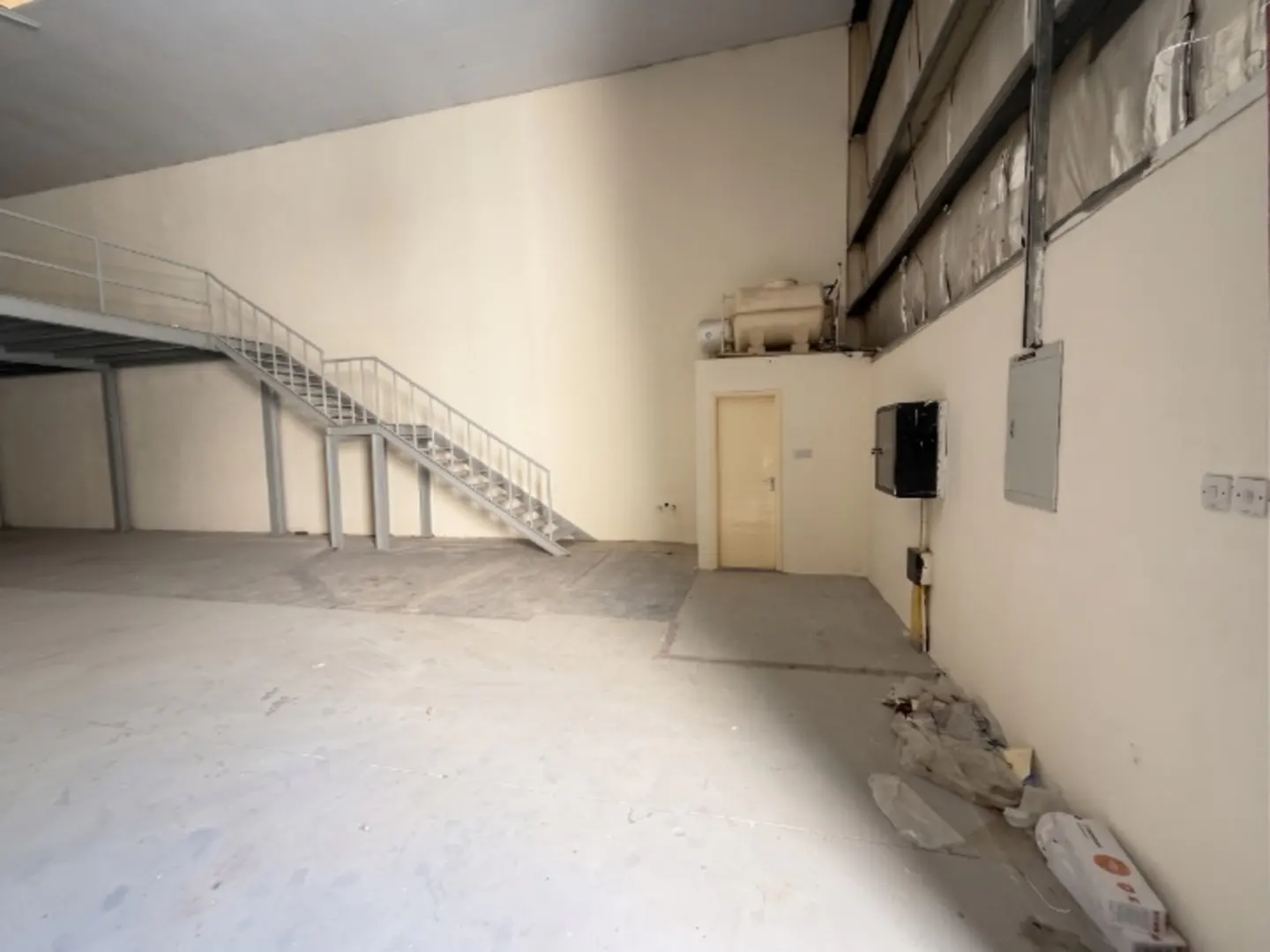 Empty warehouse interior with concrete floor, beige walls, and metal stairs leading to a mezzanine level. Electrical boxes are visible on the wall.