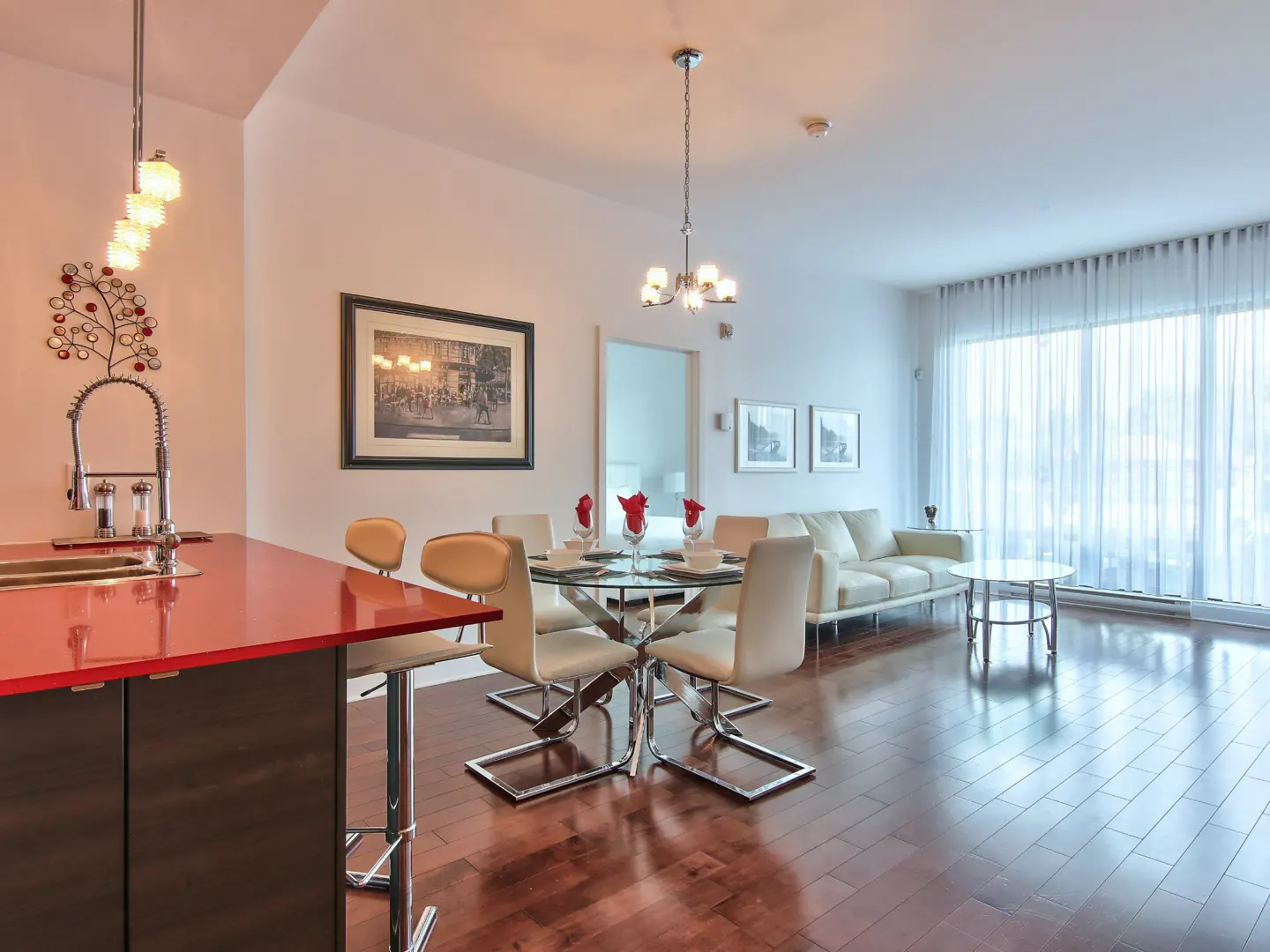 Modern open-concept living area with a red kitchen island, glass dining table, cream chairs, and a white sofa.
