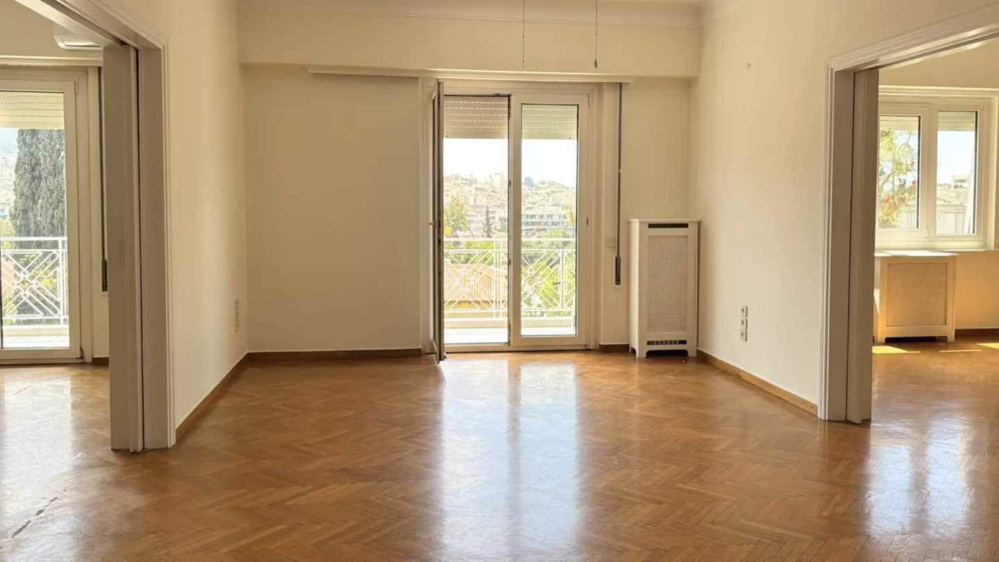 Bright, empty room with herringbone wood floors, white walls, and balcony doors with a city view.