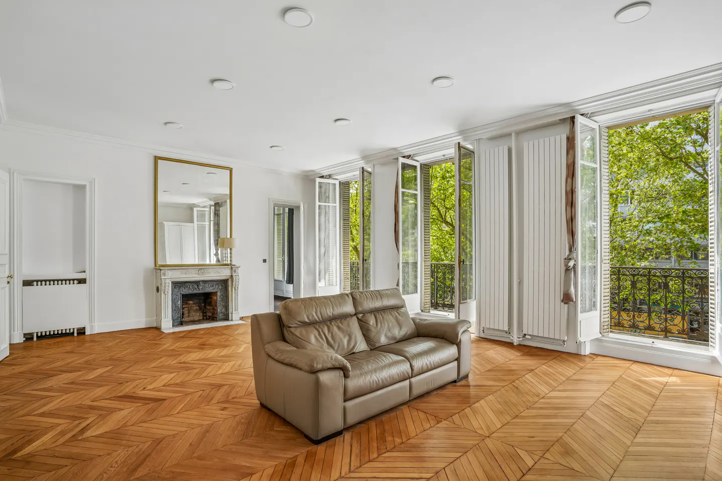 Bright living room with parquet floors, white walls, and open French doors to a balcony with green trees. A tan leather sofa sits in the center.