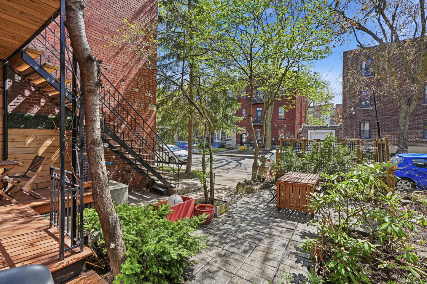 Brick building with black metal stairs, wood deck, and stone patio. Green trees and shrubs surround the outdoor space.