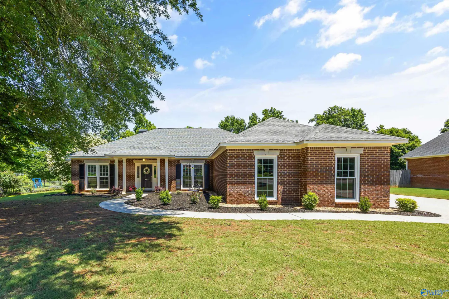 Single-story red brick house with a gray shingled roof, white trim, and a curved concrete walkway on a sunny day.