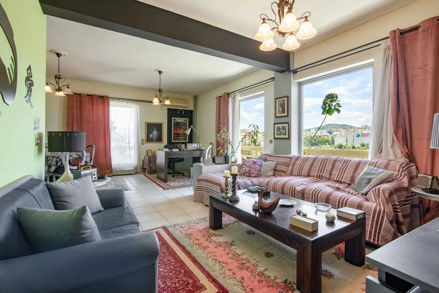 A living room with a blue sofa, striped couch, dark wood coffee table, and a desk in the background. Large windows offer a city view.