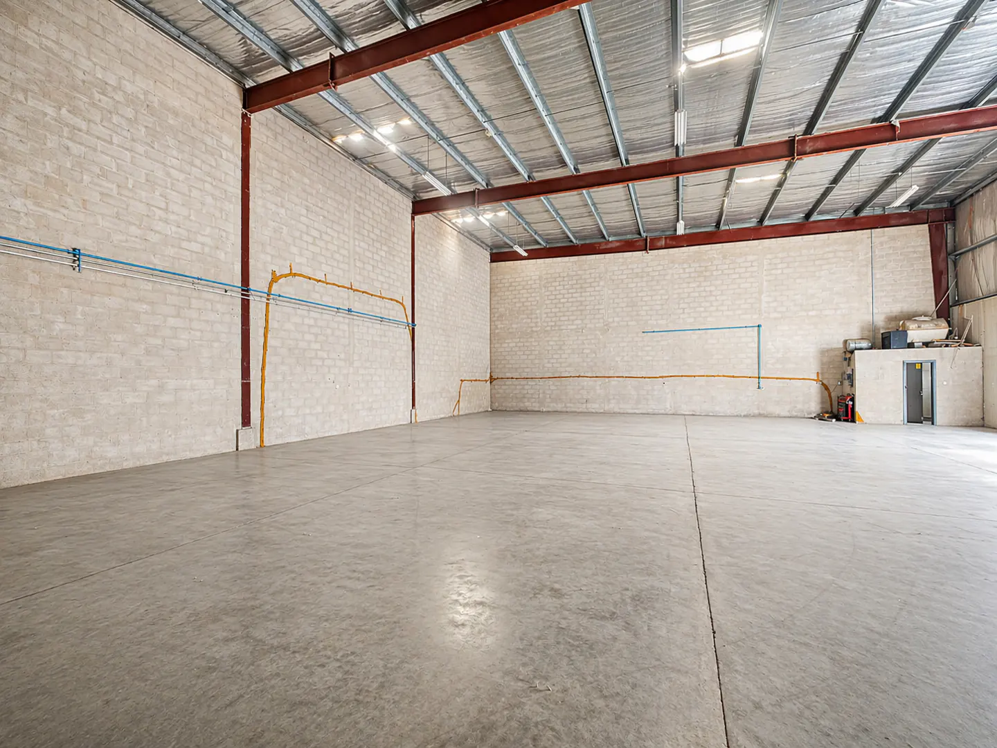 An empty warehouse with gray concrete floors, block walls, and a metal ceiling with red beams.
