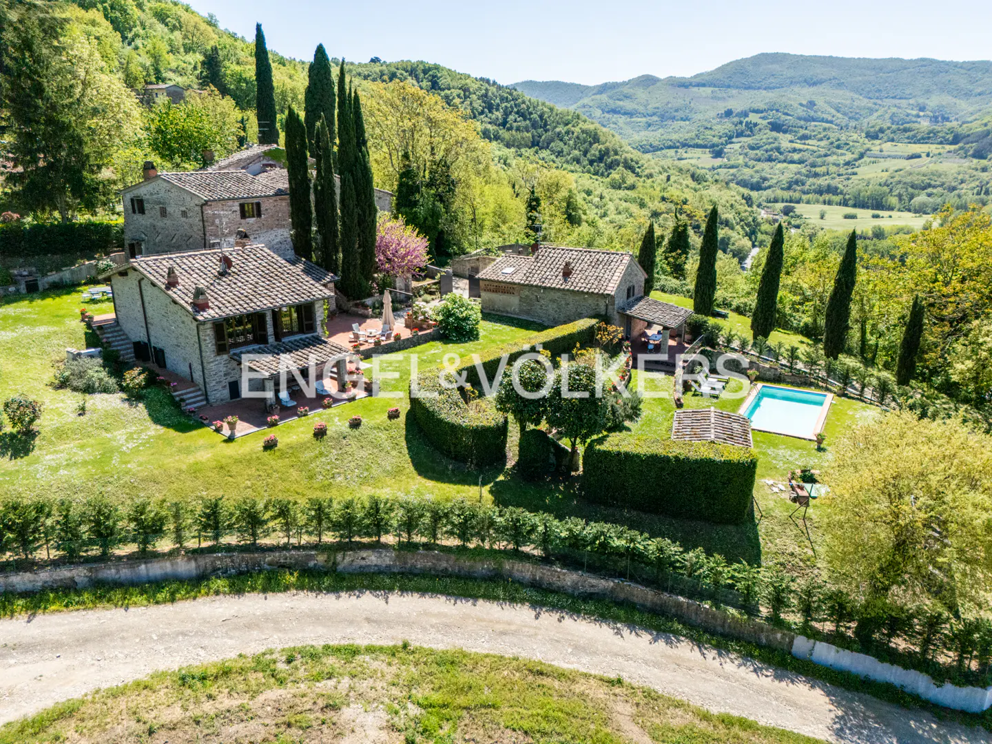 Aerial view of a stone house with a pool, surrounded by green hills and trees. The Engel & Volkers logo is visible.