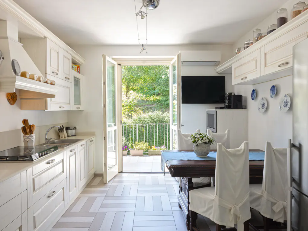 Bright kitchen with white cabinets, herringbone floors, and open doors to a balcony with green trees. A dark wood table with white-covered chairs sits nearby.