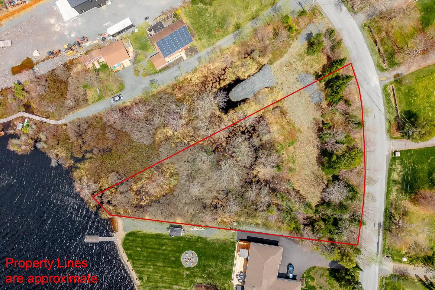 Aerial view of a triangular wooded lot next to a lake, outlined in red, with nearby houses and a road.