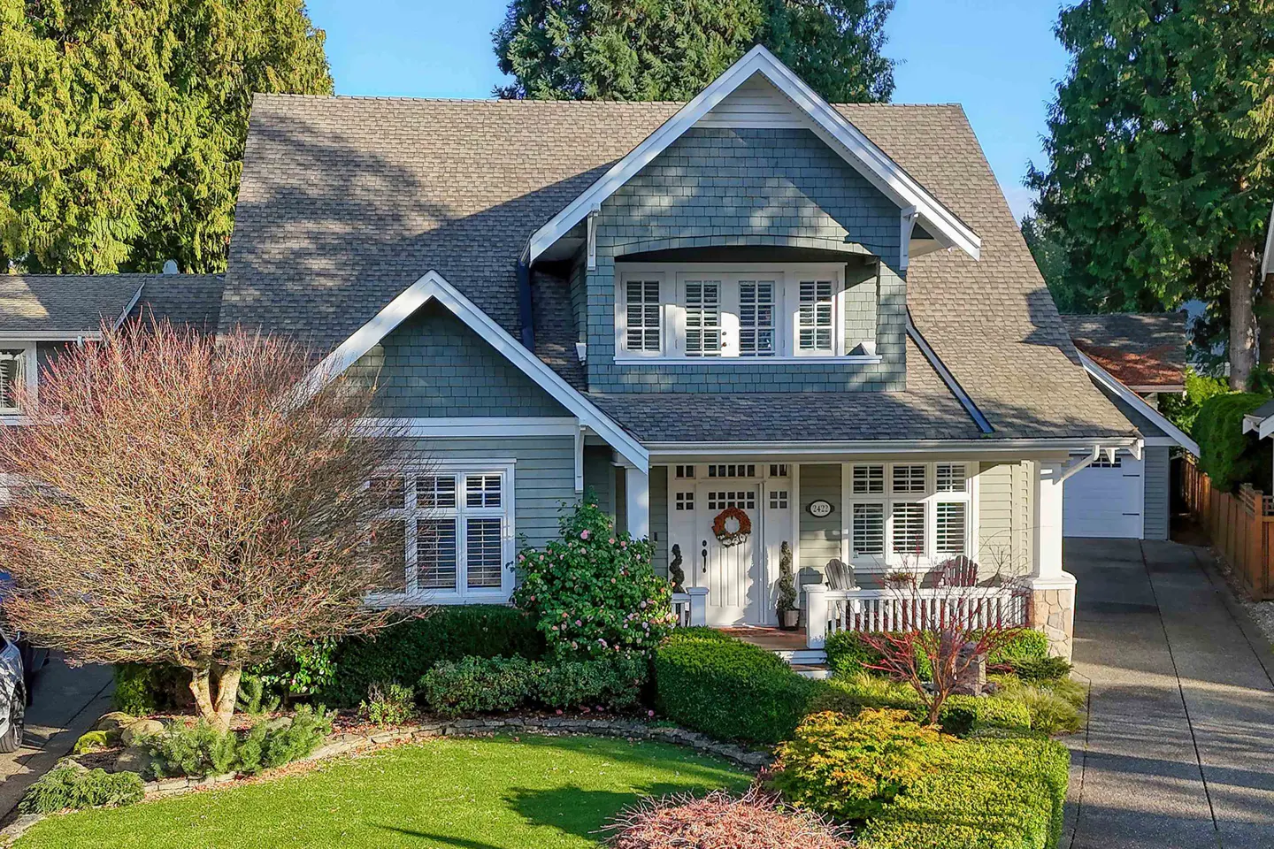 Two-story house with gray roof, green siding, white trim, and a white front door with a wreath. A green lawn and bushes are in front.