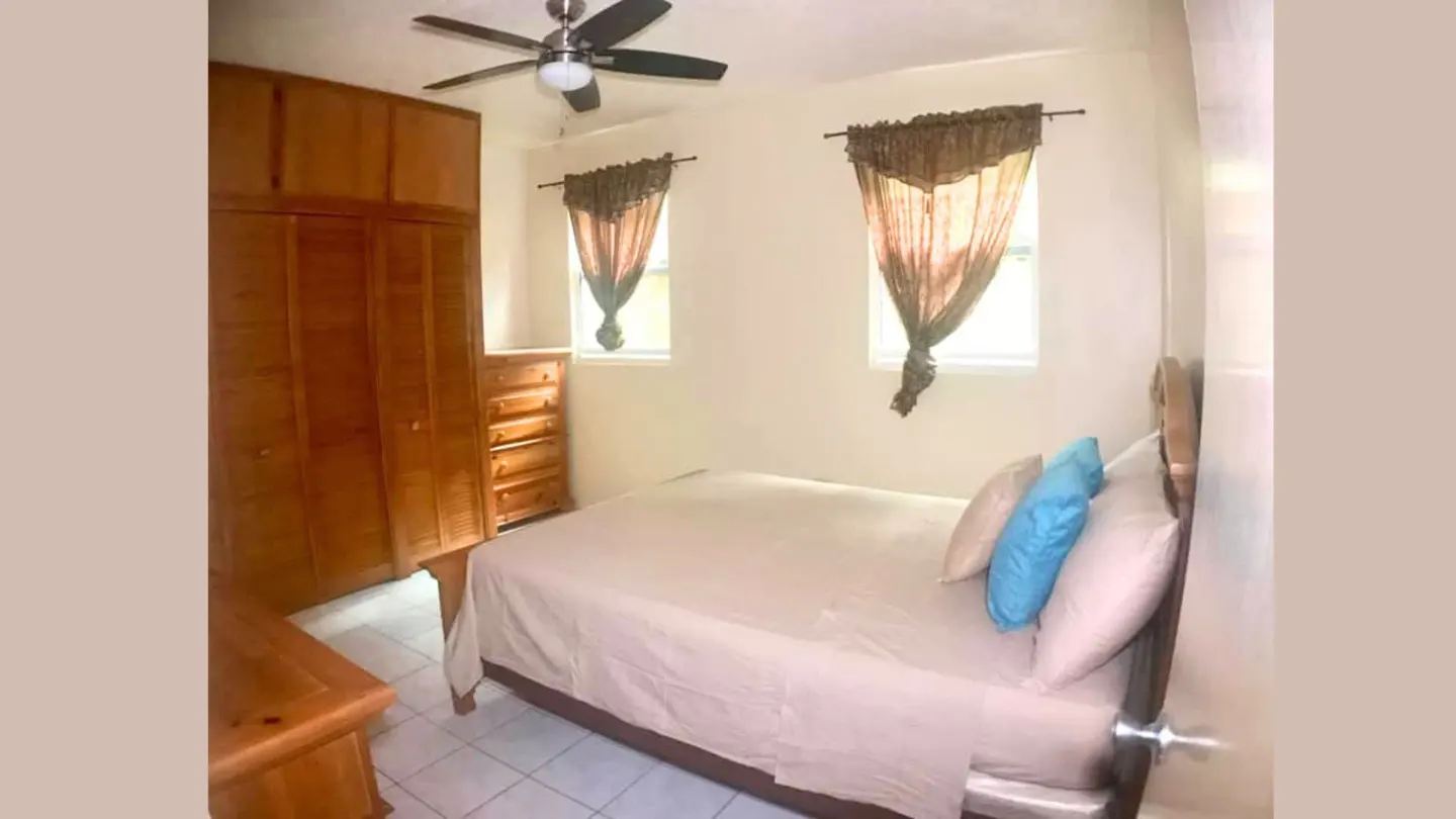 Bedroom with a bed, wooden wardrobe, dresser, ceiling fan, and two windows with brown curtains on white walls.