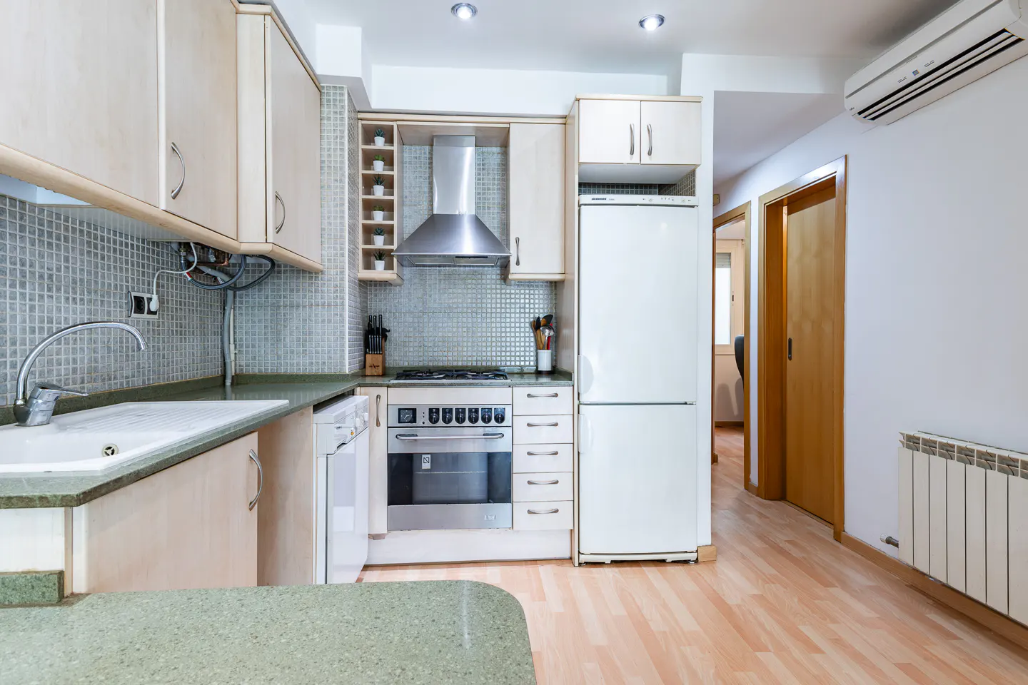 A bright kitchen with light wood cabinets, stainless steel appliances, and a green countertop. A doorway leads to another room.