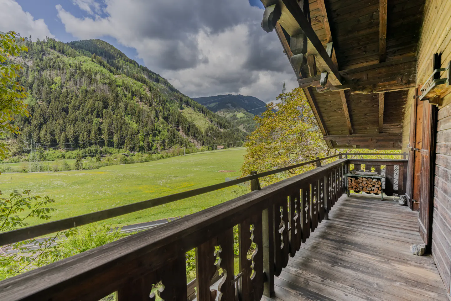 View from a wooden balcony overlooking a green valley and forested mountains. Woodpile visible on the balcony.