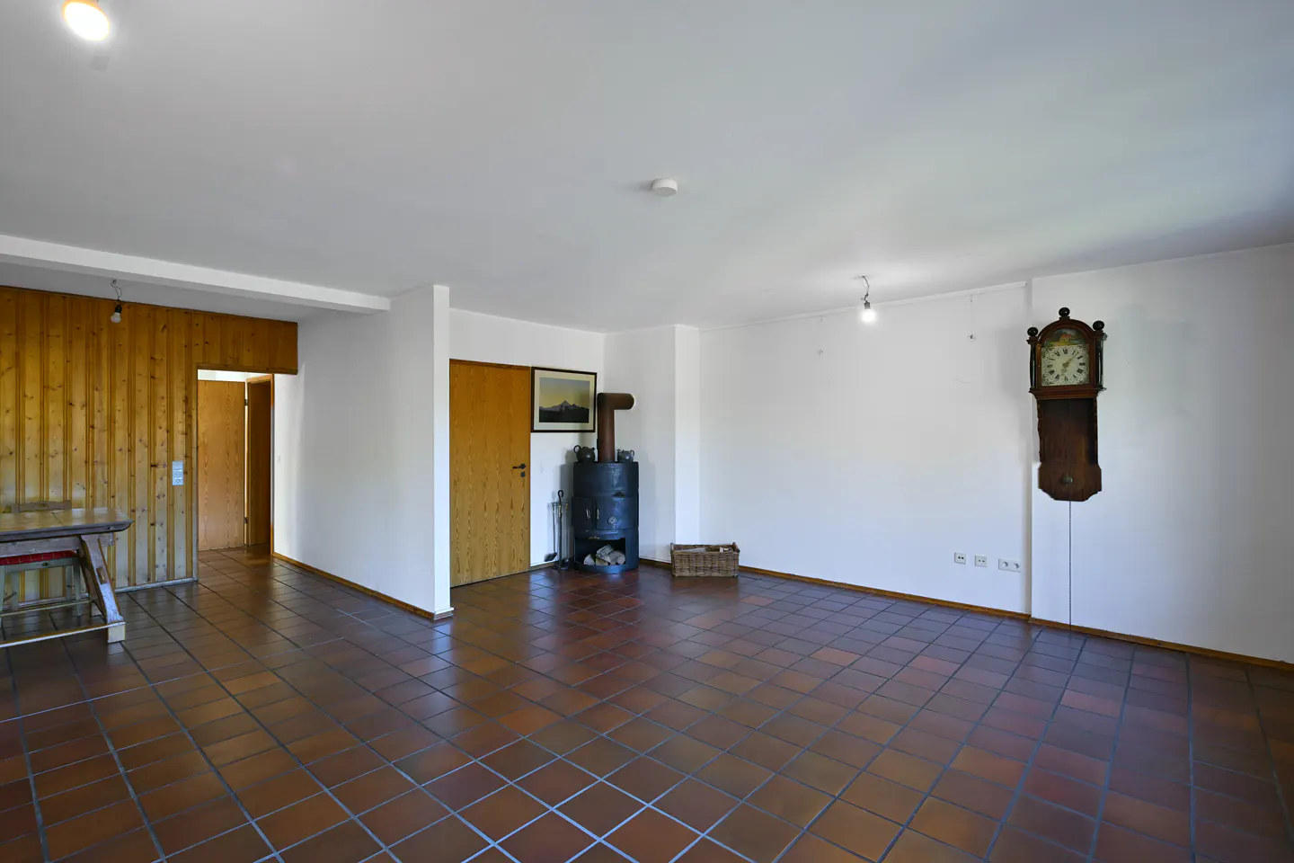 Spacious room with brown tile floor, white walls, wood paneling, and a wood-burning stove. A grandfather clock hangs on the wall.