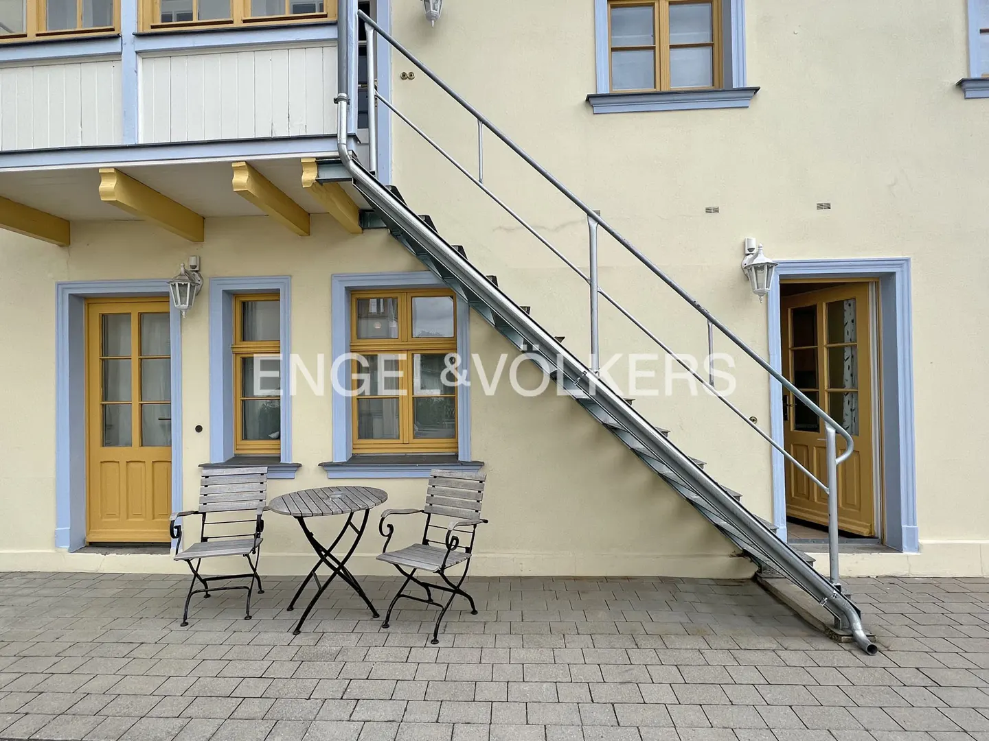 Exterior of a yellow building with blue trim, a metal staircase, and a table with two chairs.
